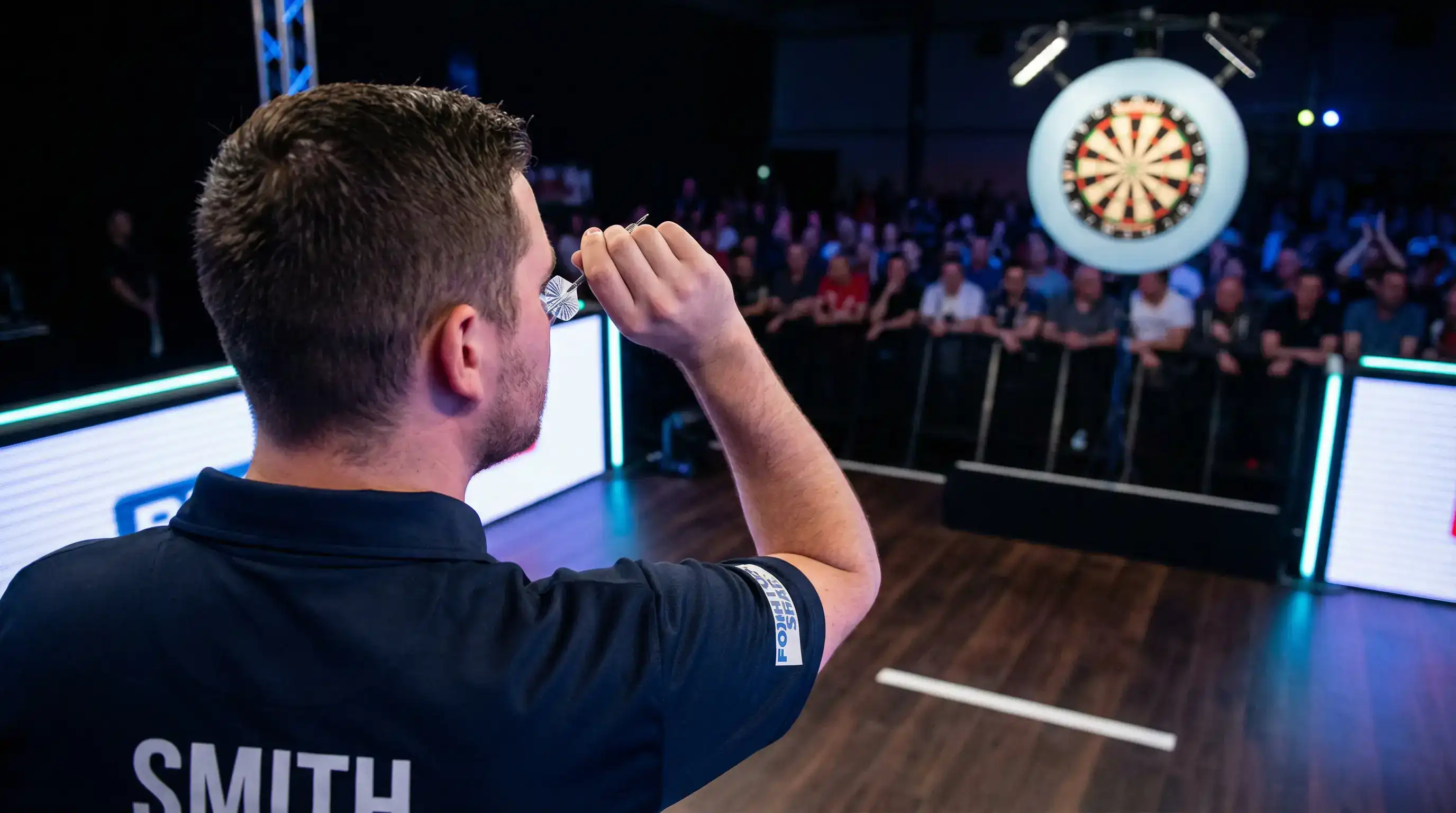 Professional darts player mid-throw on stage with dramatic spotlight illumination